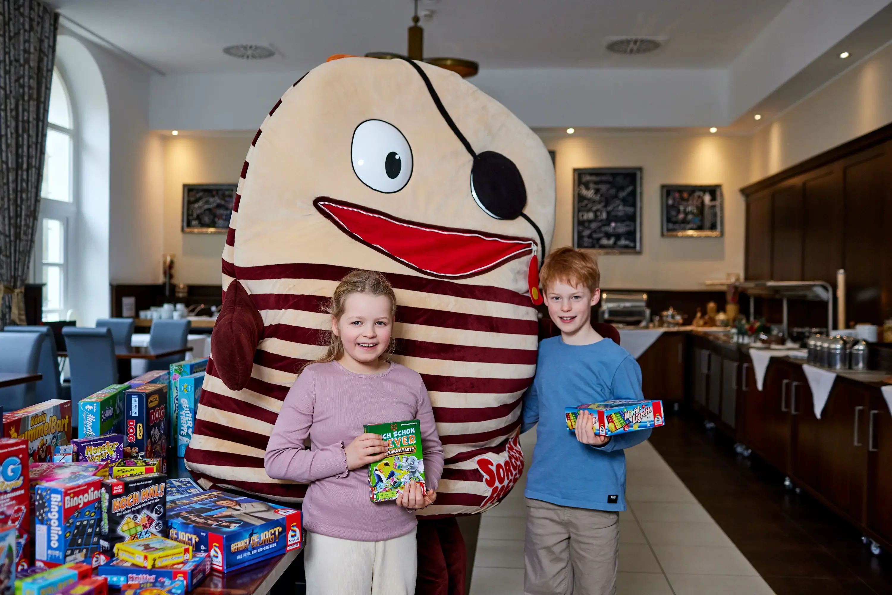 Zwei Kinder stehen in einem Raum mit Tischen und halten Spielzeugverpackungen, dahinter eine große Figur mit gestreiftem Oberkörper, Augenklappe und großem Mund.