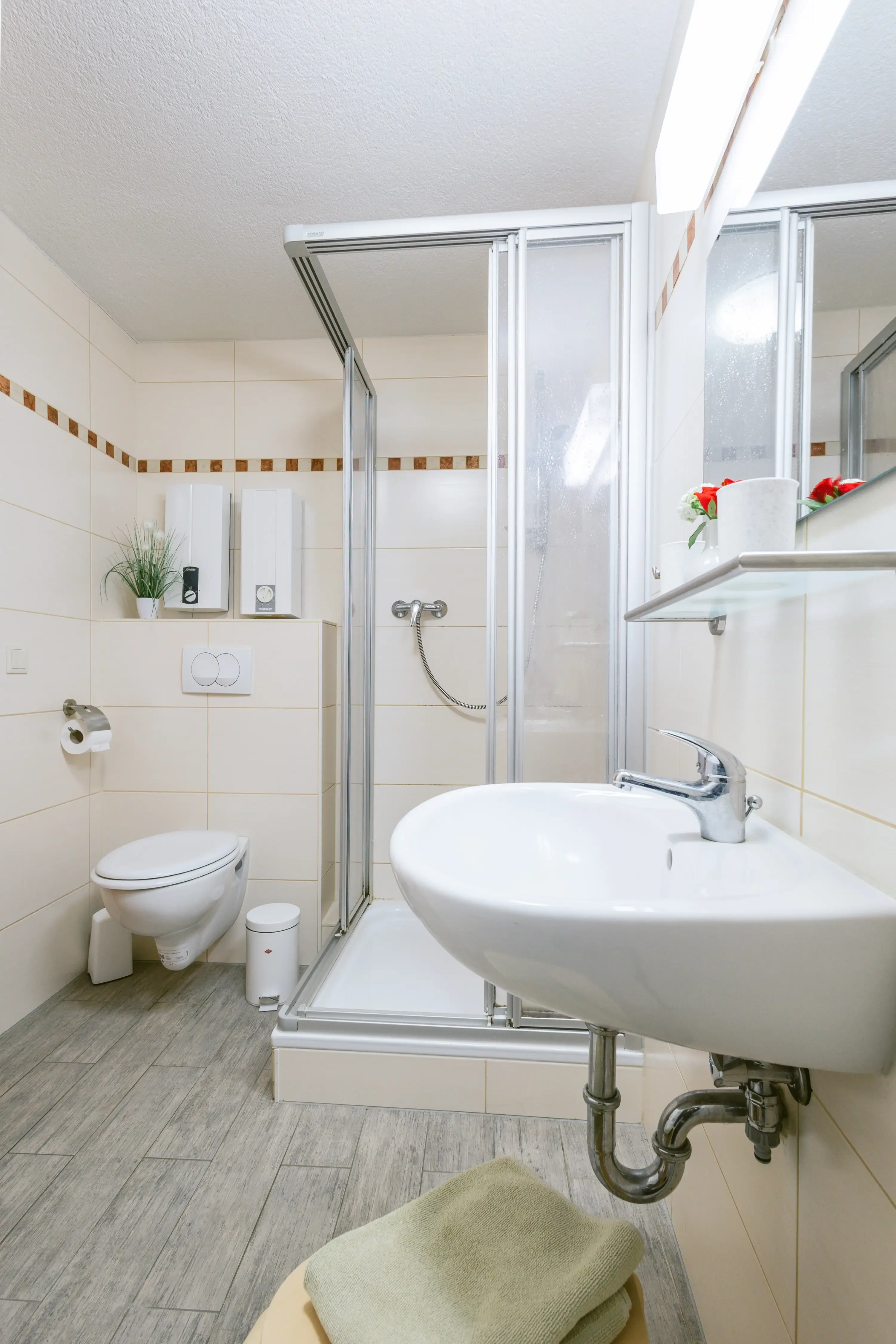 Kleines Badezimmer mit Duschkabine, Wand-WC, Waschbecken, Spiegel und Handtüchern auf dem Boden