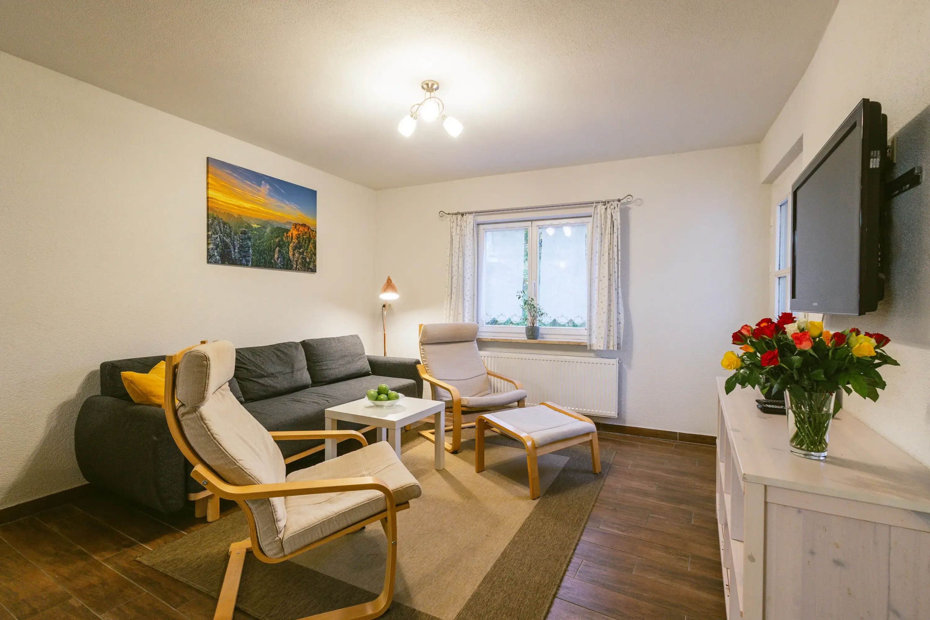 Wohnzimmer mit grauem Sofa, zwei hellen Holzsesseln mit Polstern, kleinem weißen Tisch, Wandbild über dem Sofa und Fernseher auf weißem Sideboard mit Blumenstrauß