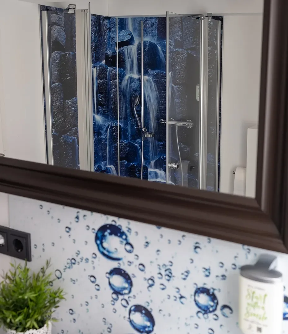 Spiegelbild einer Dusche mit Duschvorhang, der einen Wasserfall zeigt, und einem Waschbecken mit Wasserblasenmuster und einer kleinen Pflanze