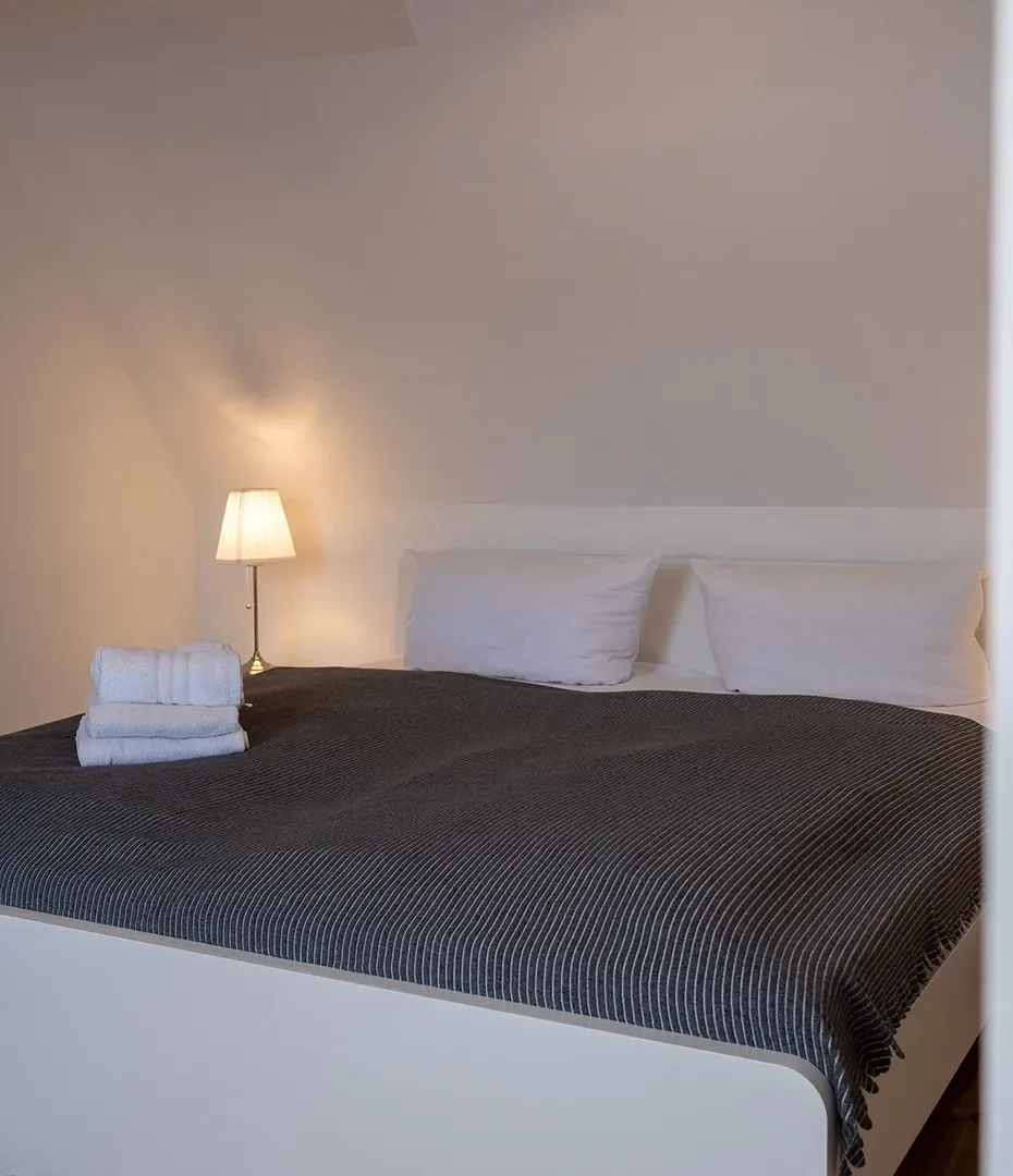 Doppelbett mit dunkler Decke, zwei weißen Kissen, Stapel weißer Handtücher und Stehlampe vor weißer Wand