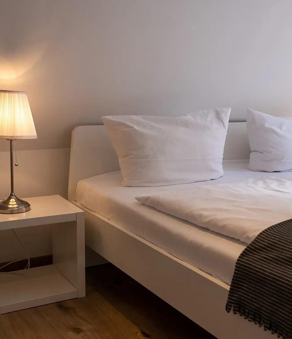 Doppelbett mit weißen Kissen und Bettdecke neben Nachttisch mit eingeschalteter Lampe in schlicht eingerichteter Ferienwohnung