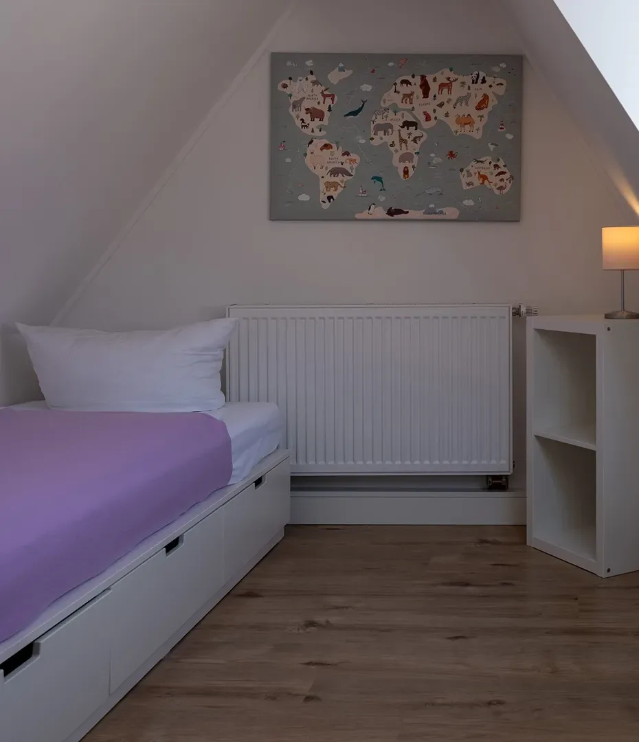 Einzelbett mit lila Bettdecke unter einer Dachschräge, darüber eine Weltkarte als Wandbild, daneben ein weißes Regal mit einer Lampe