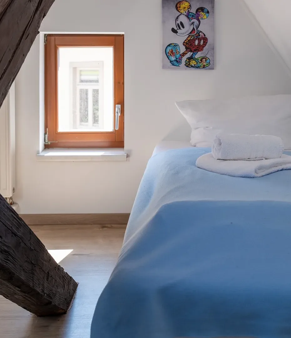 Schlafzimmer mit Einzelbett, blauem Bettbezug, Fenster mit Holzrahmen und Wandbild mit Comicfigur