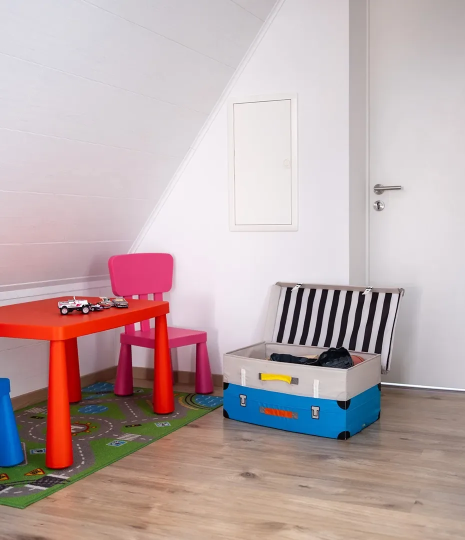 Kinderecke mit kleinem roten Tisch, pinkem Stuhl, Spielzeugautos auf dem Tisch und offenem blauen Koffer auf Holzboden unter schräger weißer Decke