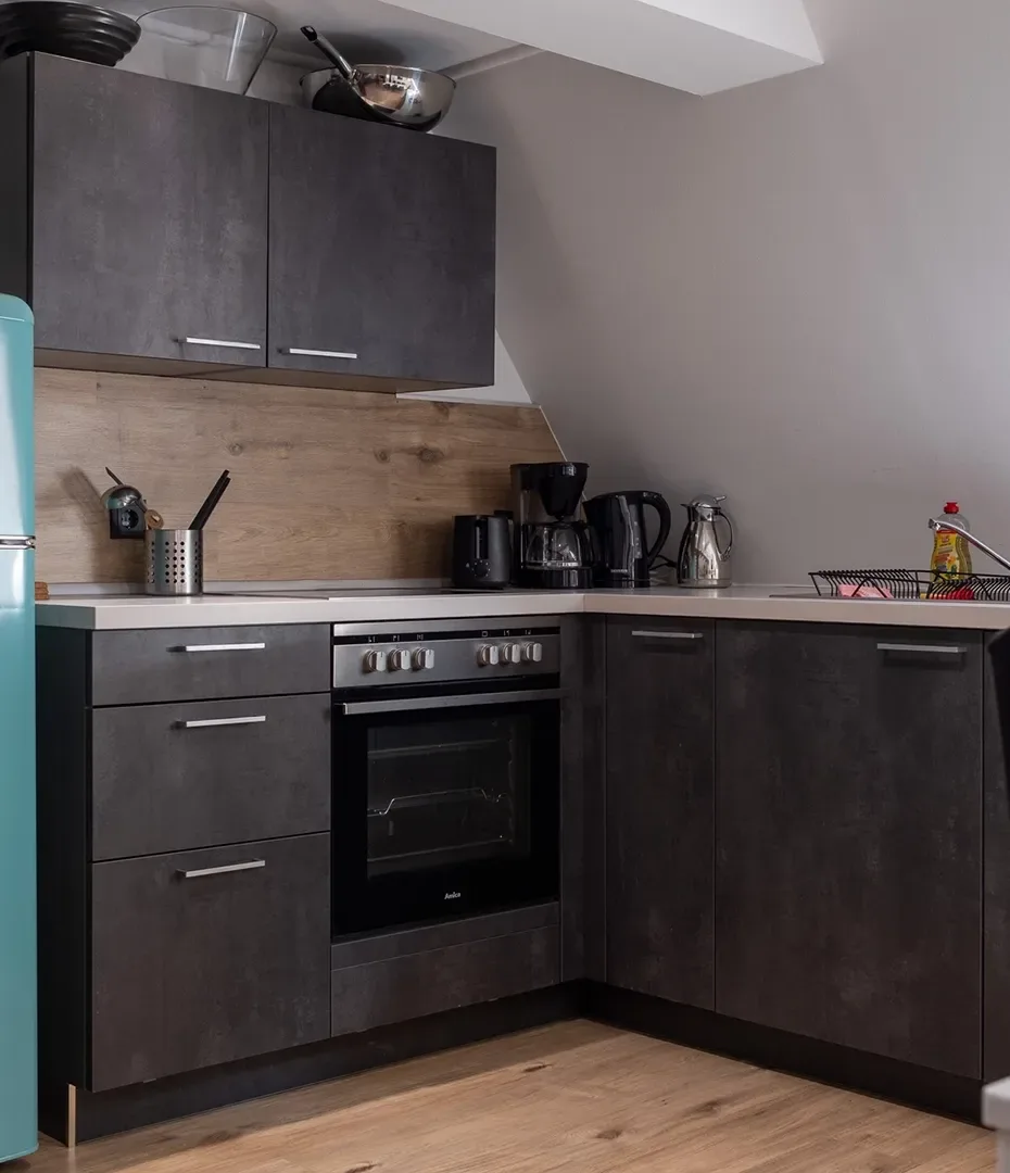 Kleine Küche mit dunklen Schränken, Backofen, Kaffeemaschine, Wasserkocher und hellblauem Kühlschrank unter einer Dachschräge