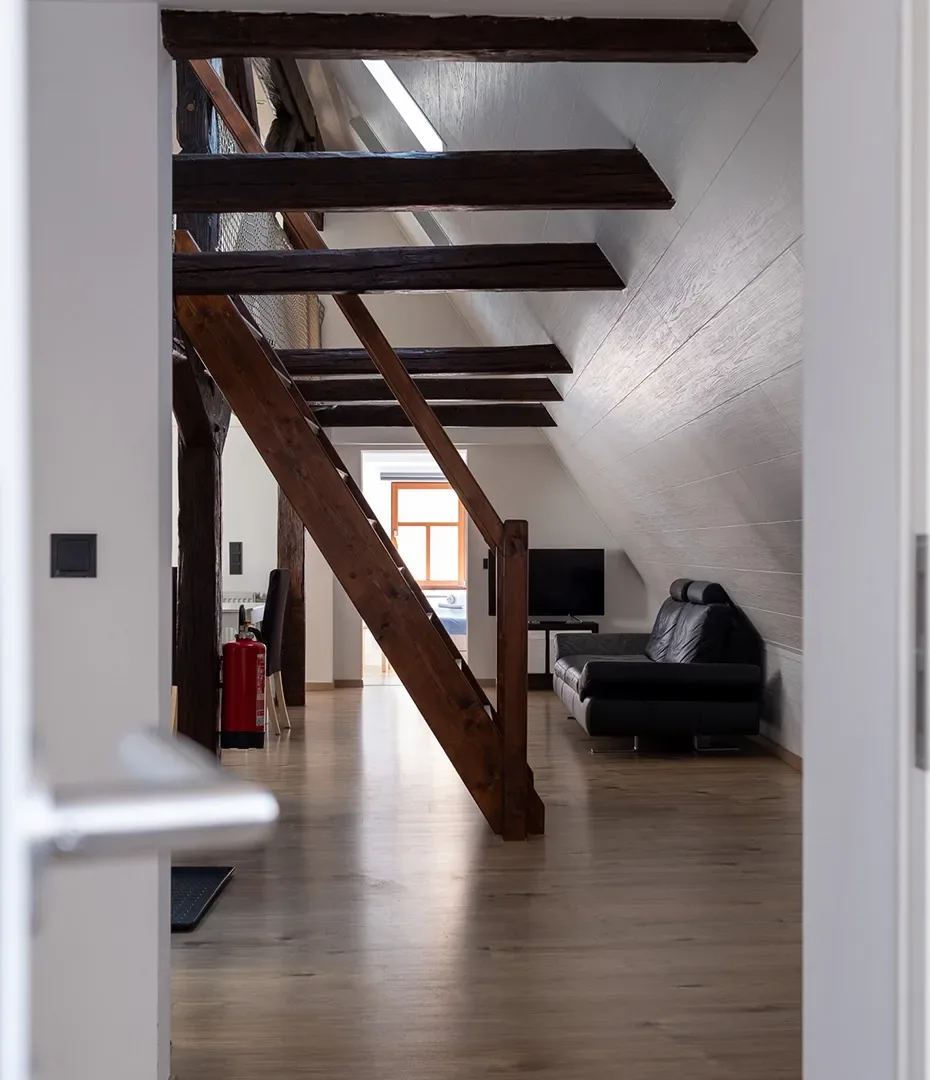 Blick durch eine geöffnete Tür in einen Dachgeschosswohnraum mit sichtbaren Holzbalken, einem schwarzen Ledersofa und einem Fernseher