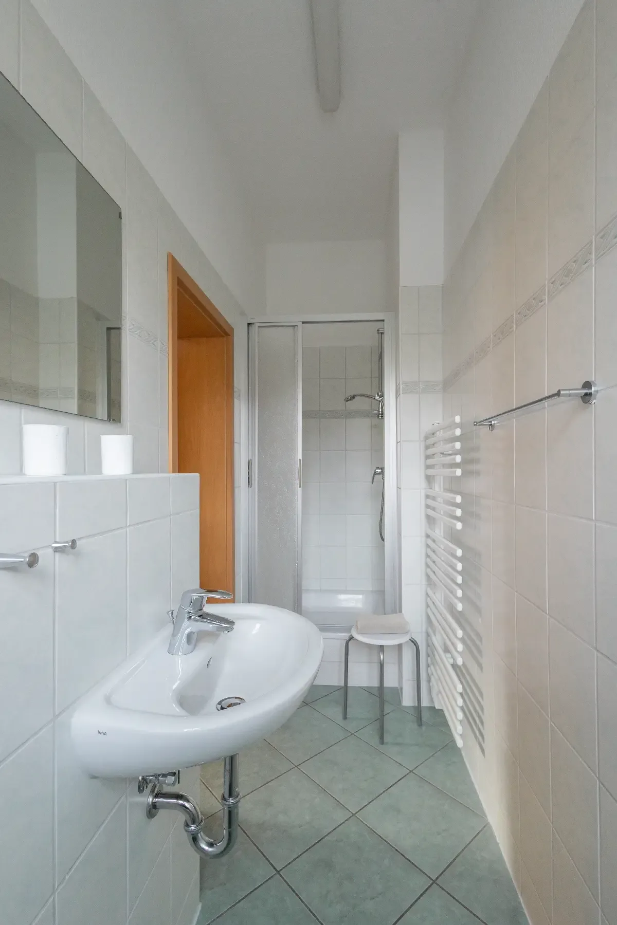 Kleines Badezimmer mit weißem Waschbecken, Spiegel, Duschkabine, Handtuchheizkörper und Hocker