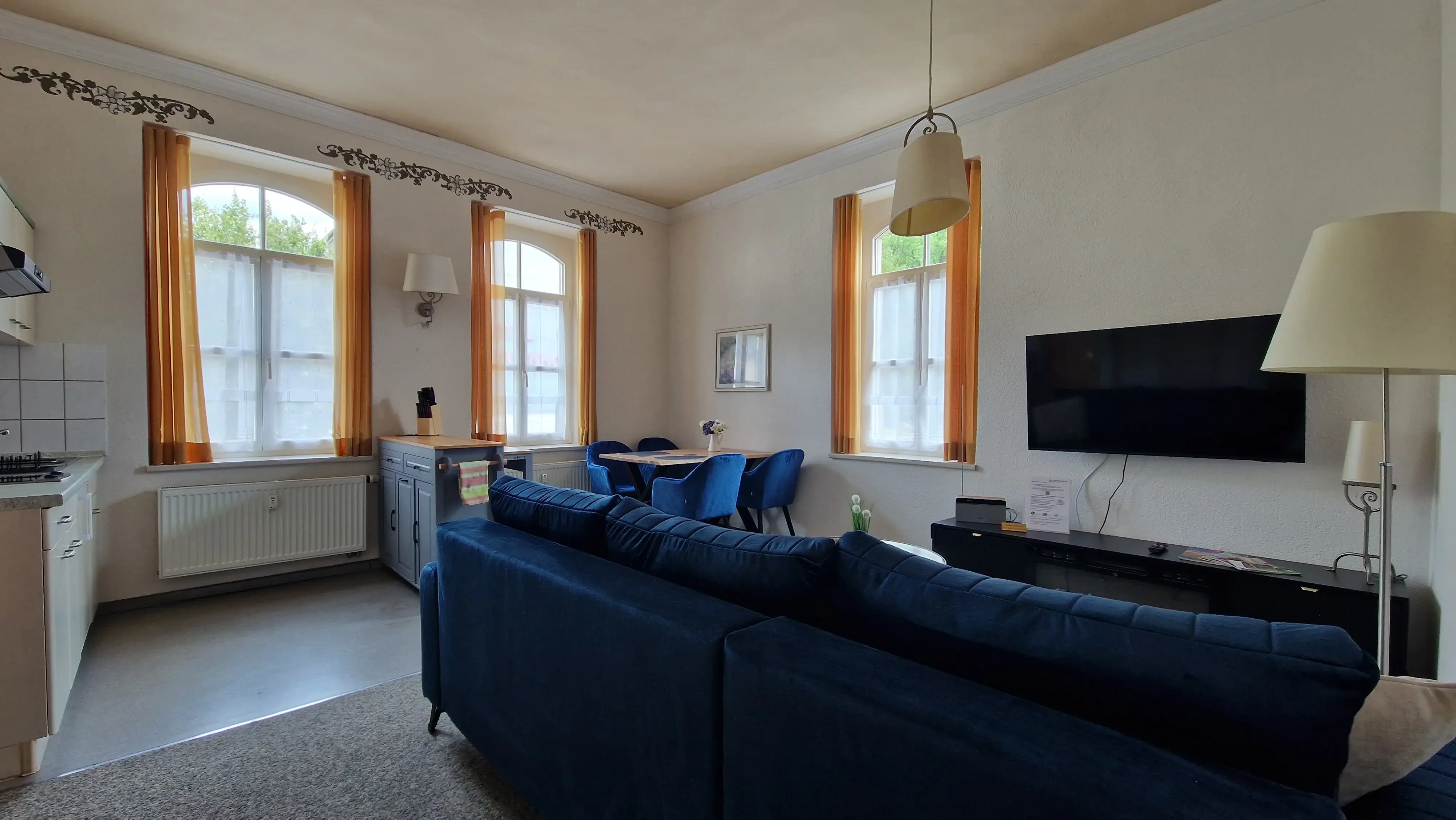 Wohnzimmer mit blauer Couch, Esstisch mit blauen Stühlen, zwei Fenstern mit gelben Vorhängen und Wandfernseher