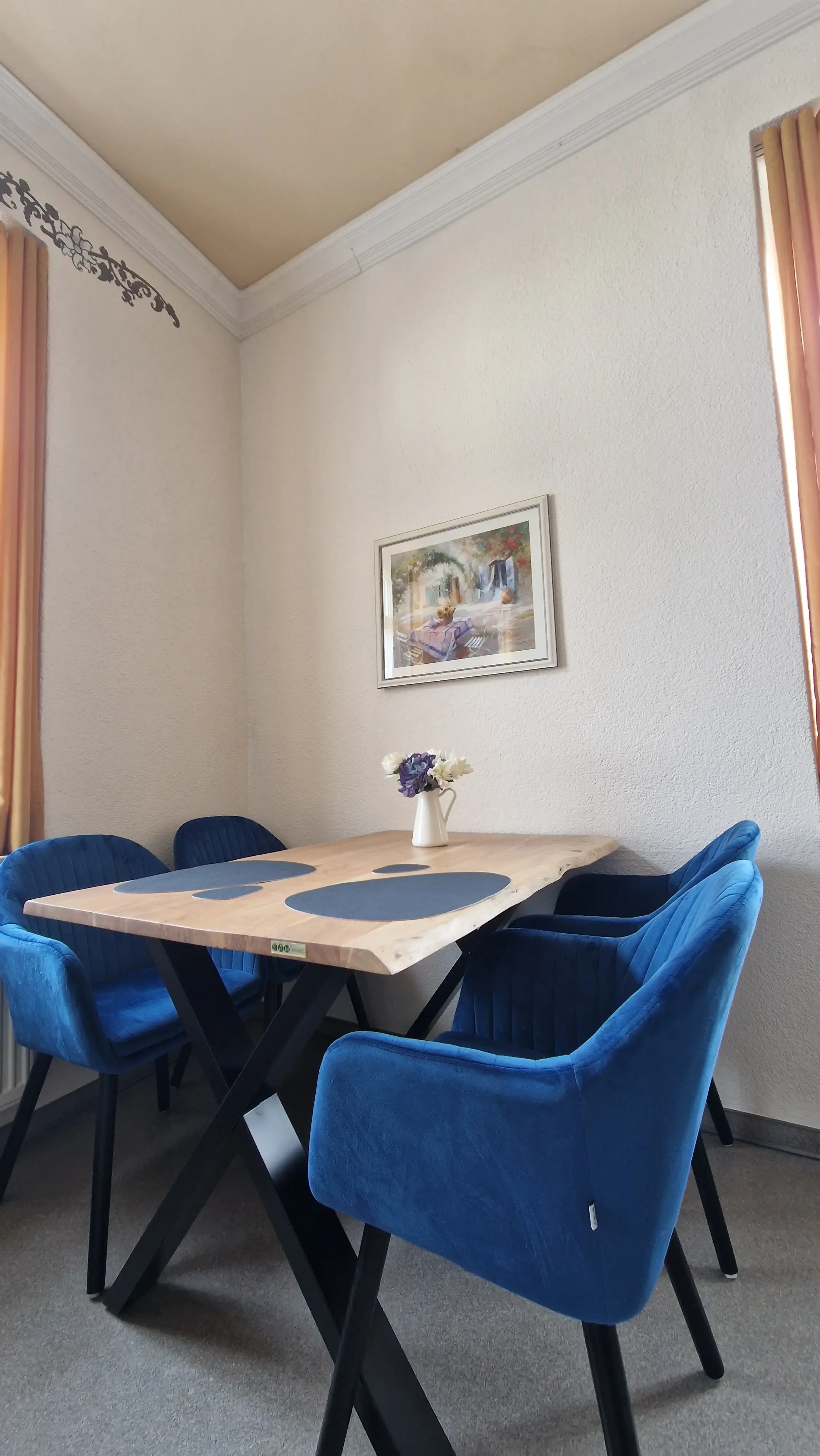 Holztisch mit schwarzer Tischdecke und vier blauen gepolsterten Stühlen in einer hellen Ecke mit Bild an der Wand