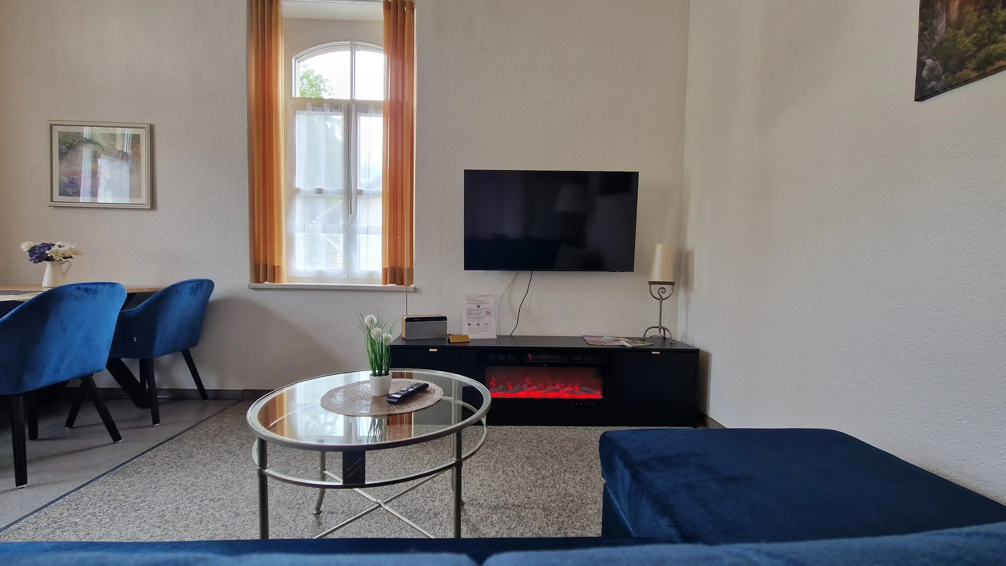 Wohnbereich mit blauem Sofa, rundem Glastisch, Wandfernseher und Esstisch mit blauen Stühlen vor Fenster