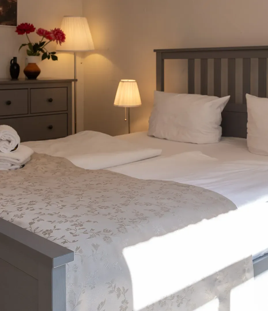 Doppelbett mit weißen Kissen und Decke vor grauem Kopfteil neben Kommode mit Blumen und zwei Lampen