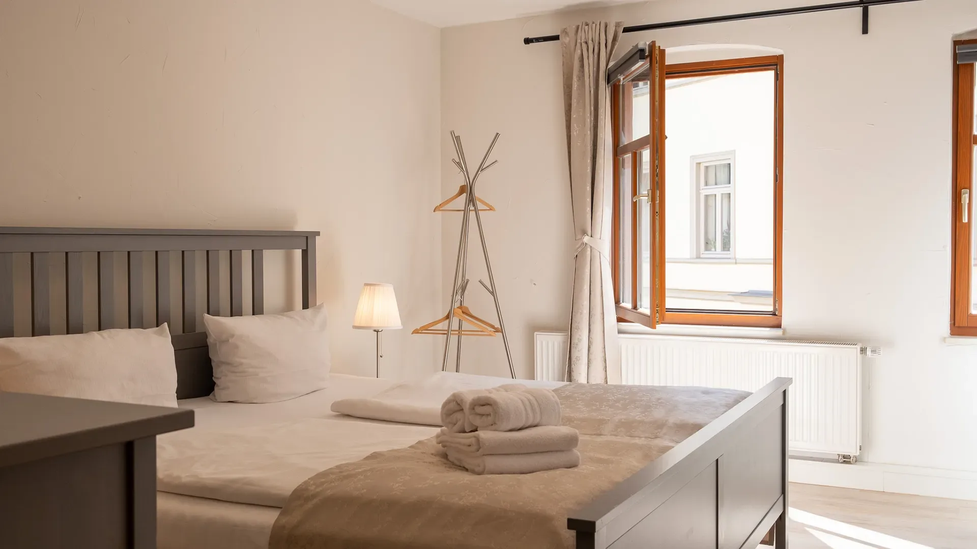 Schlafzimmer mit Doppelbett, offenem Fenster, Gardinen, Stehlampe und auf dem Bett gestapelten Handtüchern