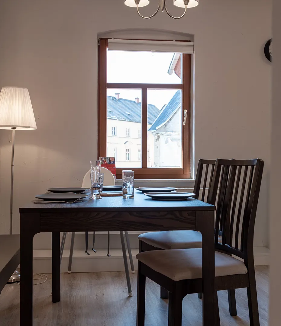 Essbereich mit dunklem Holztisch, vier Stühlen mit hellen Sitzpolstern, Stehlampe und Fenster mit Blick auf Nachbargebäude