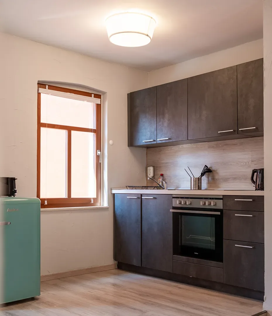 Kleine Küche mit dunklen Oberschränken, Herd mit Backofen, hellem Fußboden, Fenster mit Holzrahmen und mintgrünem Kühlschrank