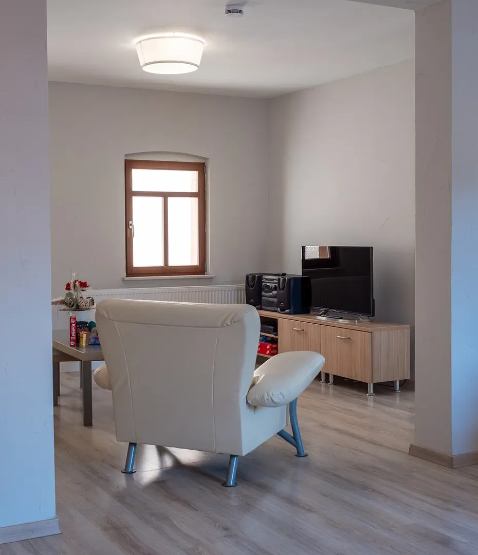 Wohnzimmer mit hellen Wänden, einem Fenster, einem weißen Sessel, einem Fernseher auf einem Sideboard und einem kleinen Tisch mit Dekoration