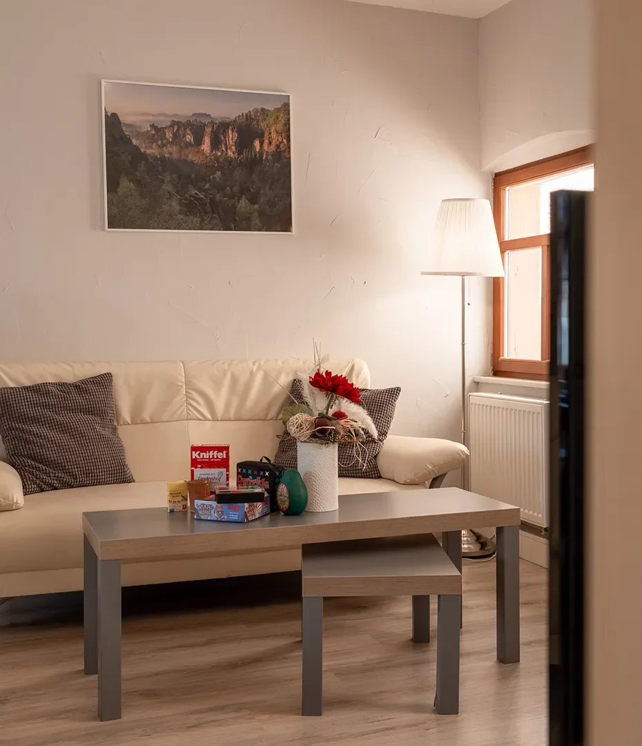 Wohnzimmer mit hellbeiger Couch, zwei Kissen, grauem Couchtisch mit Spielen und Dekoration, Stehlampe und Fenster mit Holzrahmen