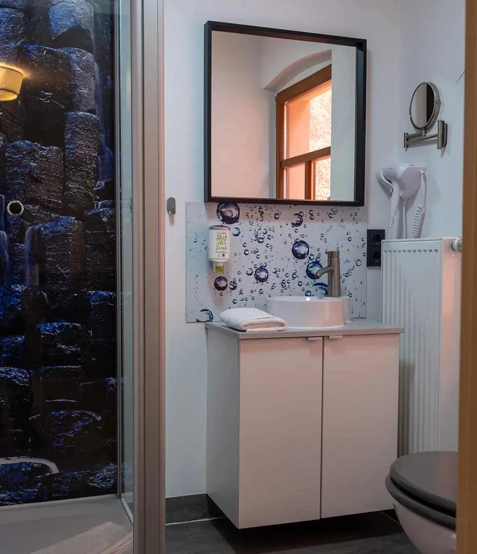 Kleines Badezimmer mit weißem Waschbeckenunterschrank, rundem Waschbecken, Spiegel, Duschkabine mit dunkler Steinwand und Handtuchhalter am Heizkörper