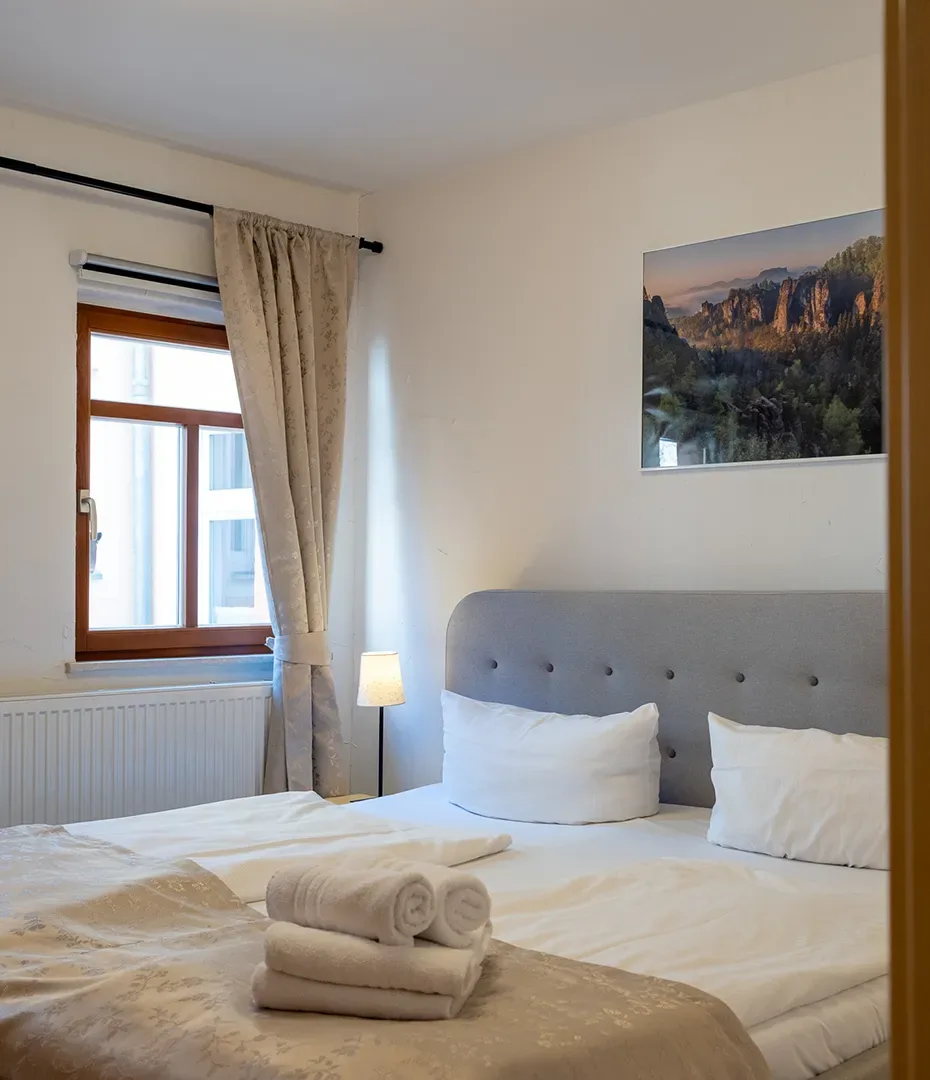Schlafzimmer mit Doppelbett, grauem gepolstertem Kopfteil, weißen Kissen und Bettwäsche, zwei aufgerollten Handtüchern auf der Bettdecke, Fenster mit Vorhang und Stehlampe neben dem Bett