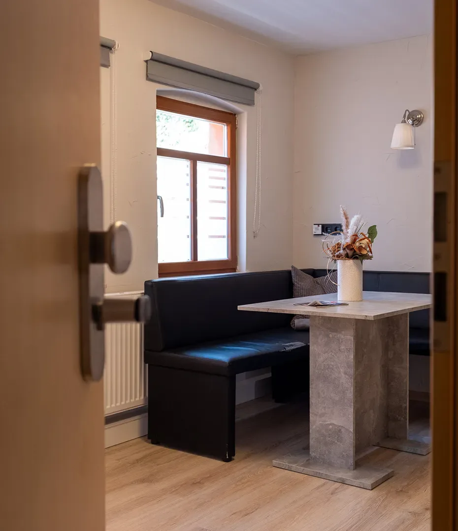 Blick durch geöffnete Tür auf kleine Essecke mit schwarzer Eckbank, quadratischem Steintisch und Blumendekoration vor Fenster