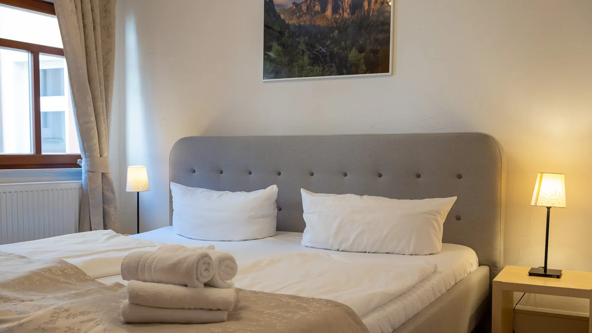 Hell eingerichtetes Schlafzimmer mit Doppelbett, zwei weißen Kissen, grauem gepolstertem Kopfteil, zwei Nachttischen mit Lampen und Fenster mit Vorhängen