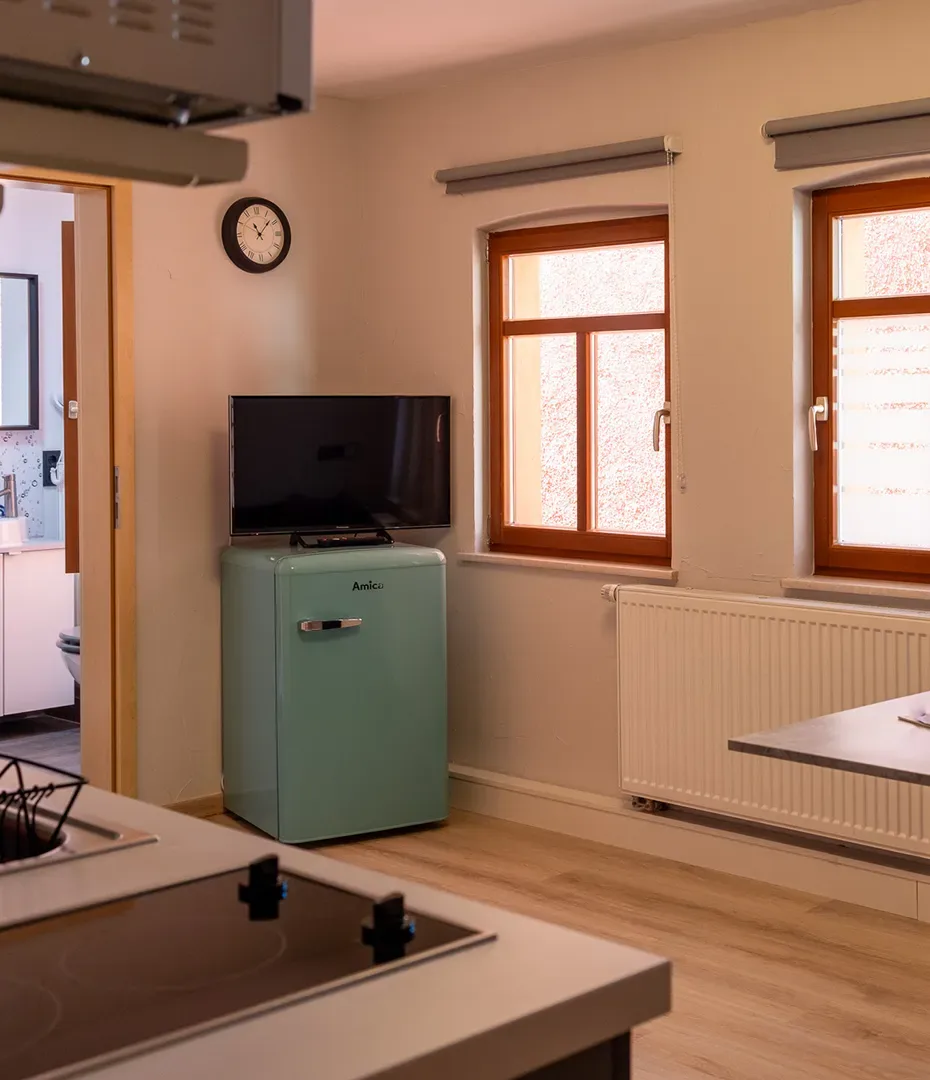 Kleine Küche mit kleinem türkisfarbenem Kühlschrank, Fernseher darauf, zwei Fenstern mit Rollos und Wanduhr