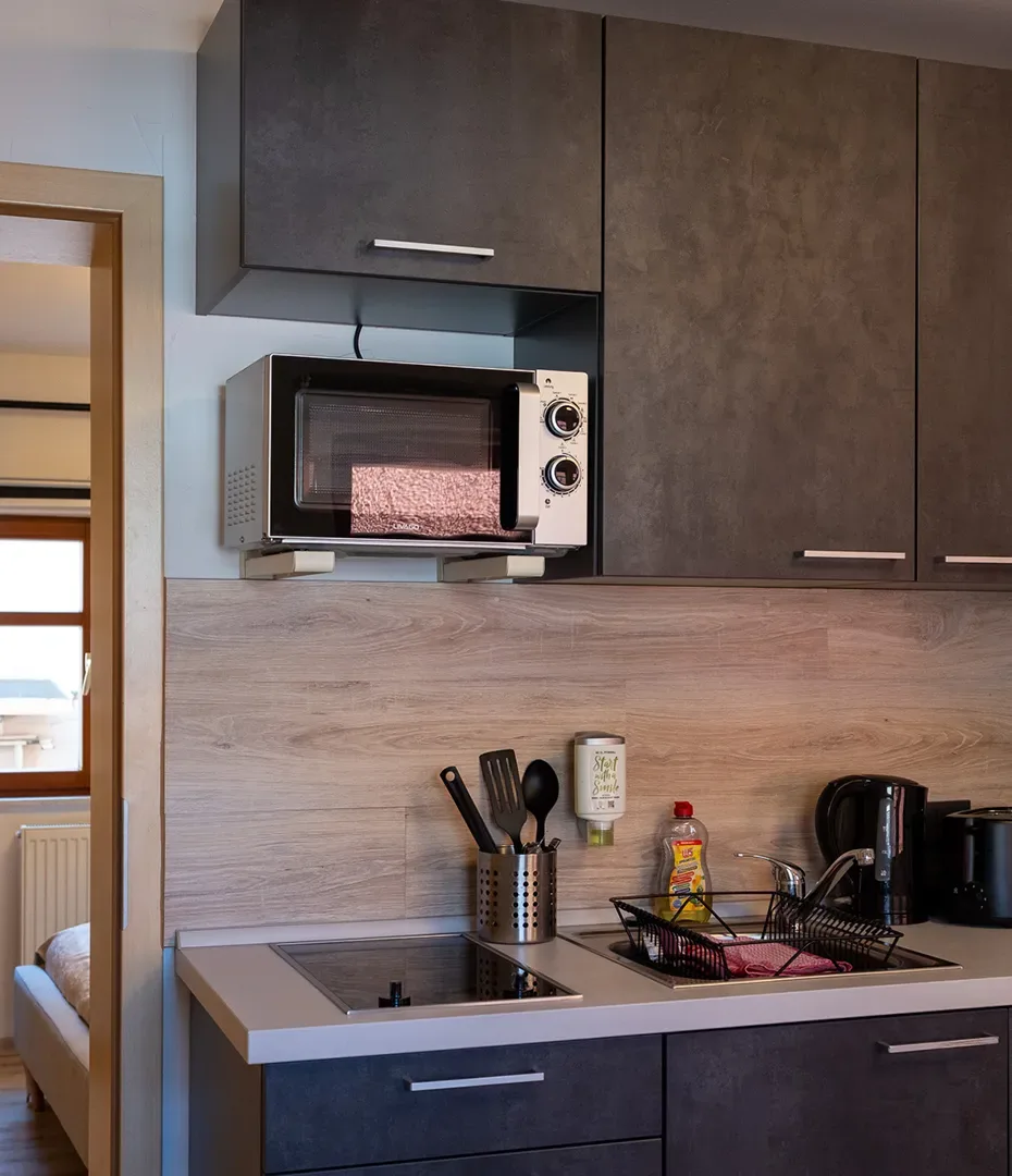 Kleine Küche mit dunklen Oberschränken, Mikrowelle, Kochfeld, Spüle, Geschirrkorb und Besteckbehälter