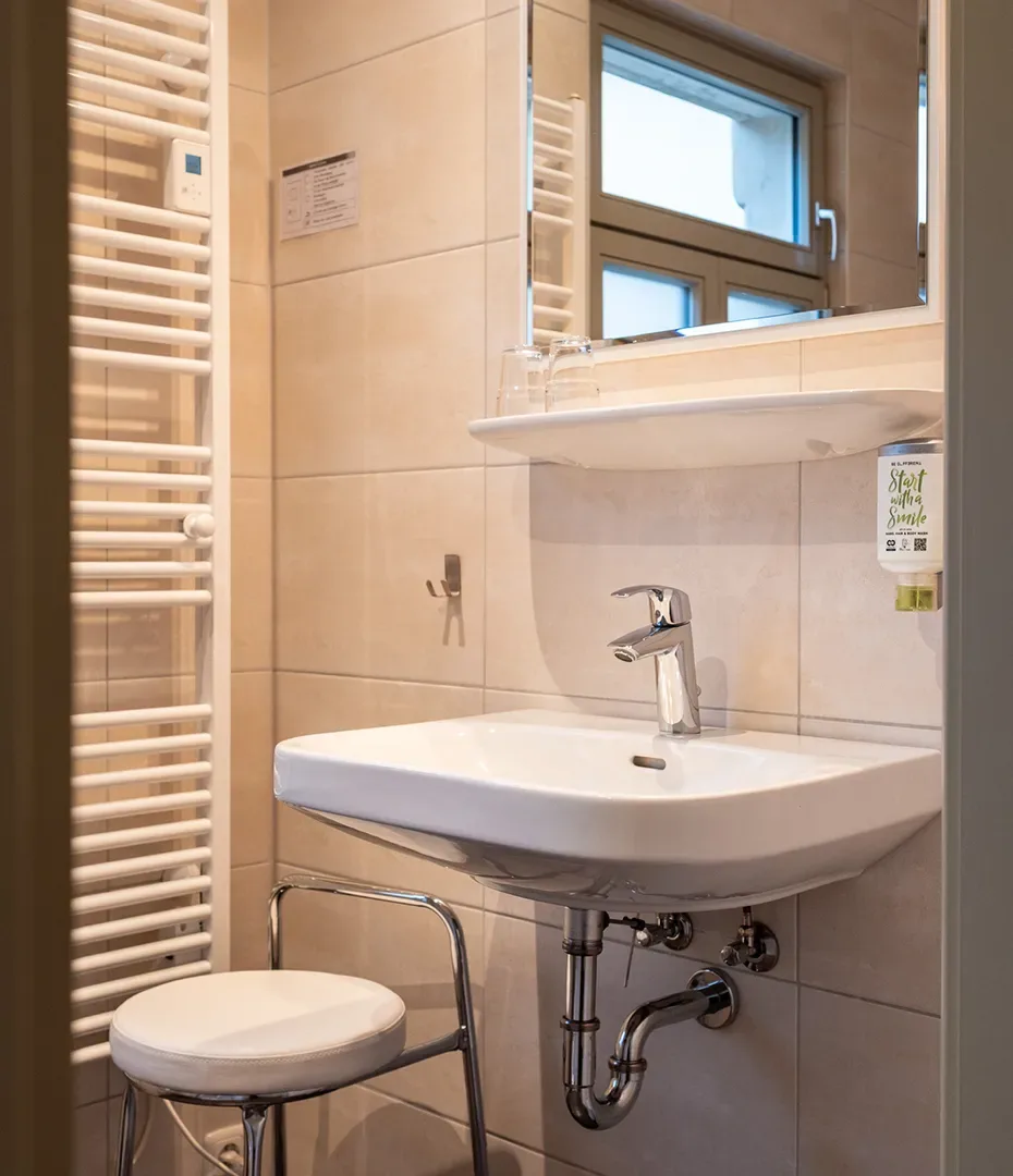 Waschbecken mit Armatur, Spiegel, zwei Gläser auf dem Waschbeckenrand, Hocker mit weißem Sitz und Handtuchheizkörper an der Wand in einem Badezimmer