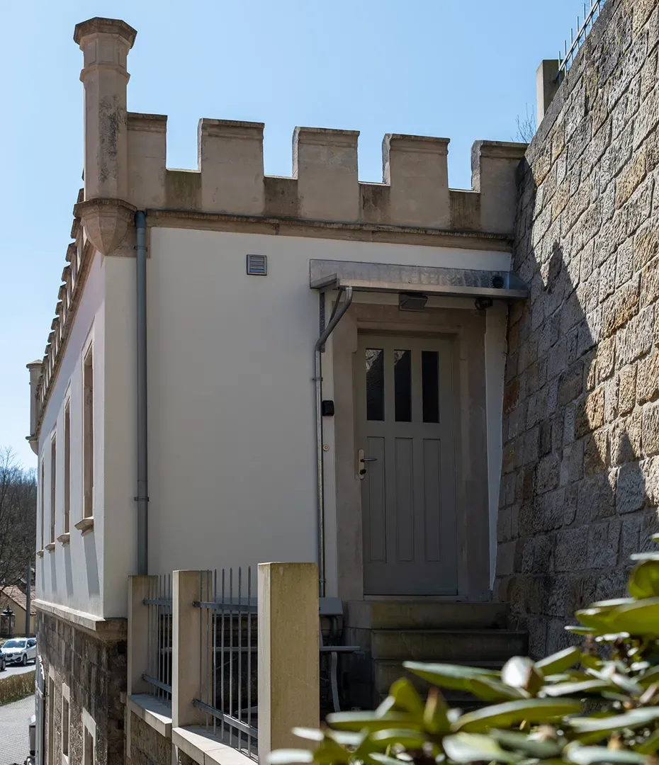 Eingang eines Gebäudes mit hellgrauer Tür, Treppe und Zinnenmauerwerk auf dem Dach