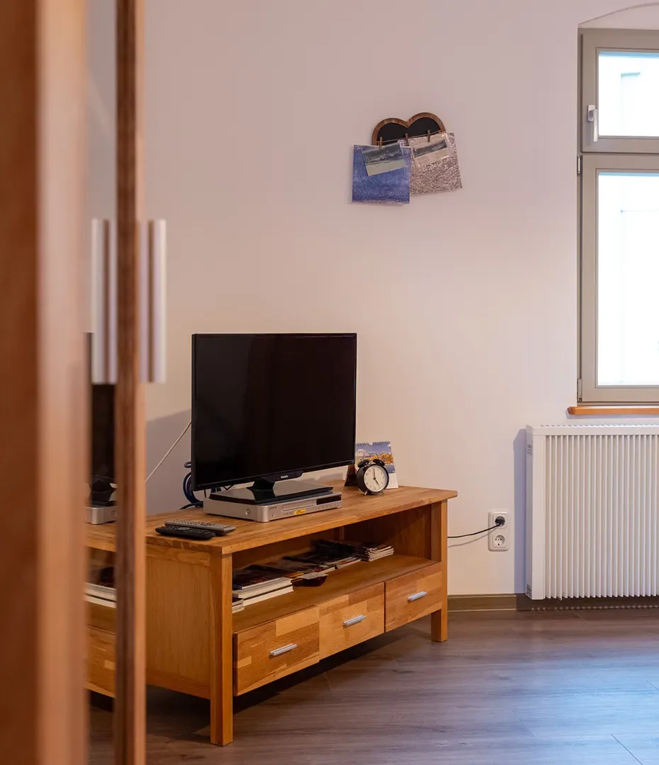 Holz-TV-Möbel mit Flachbildfernseher, Wecker und Fernbedienung vor weißer Wand mit zwei an Pinnwand befestigten Postkarten neben Fenster und Heizkörper