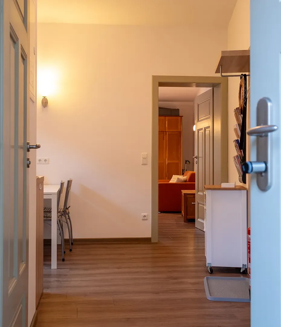 Flur mit Holzboden, links ein Tisch mit Stühlen, rechts ein Regal und Blick in ein Wohnzimmer mit Sofa und Schrank