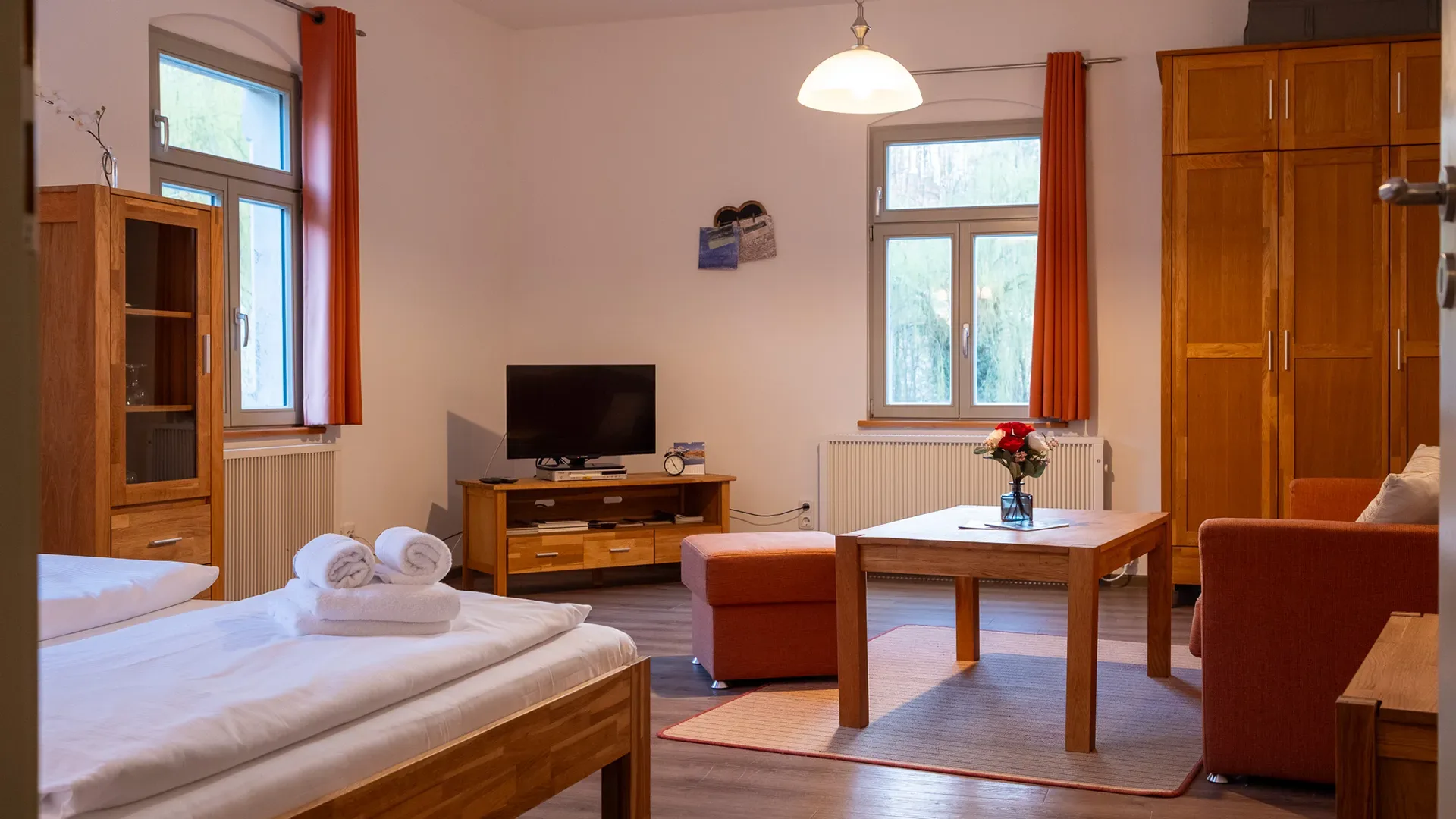 Wohnzimmer mit Holzmöbeln, Sofa, Fernseher, Tisch mit Blumenvase und zwei Fenstern mit Vorhängen