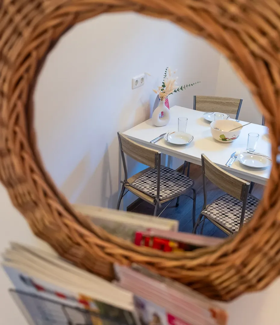 Essplatz mit weißem Tisch, drei Stühlen mit gemusterten Sitzflächen und einer Vase mit Blumen, sichtbar durch einen runden Korbrahmen