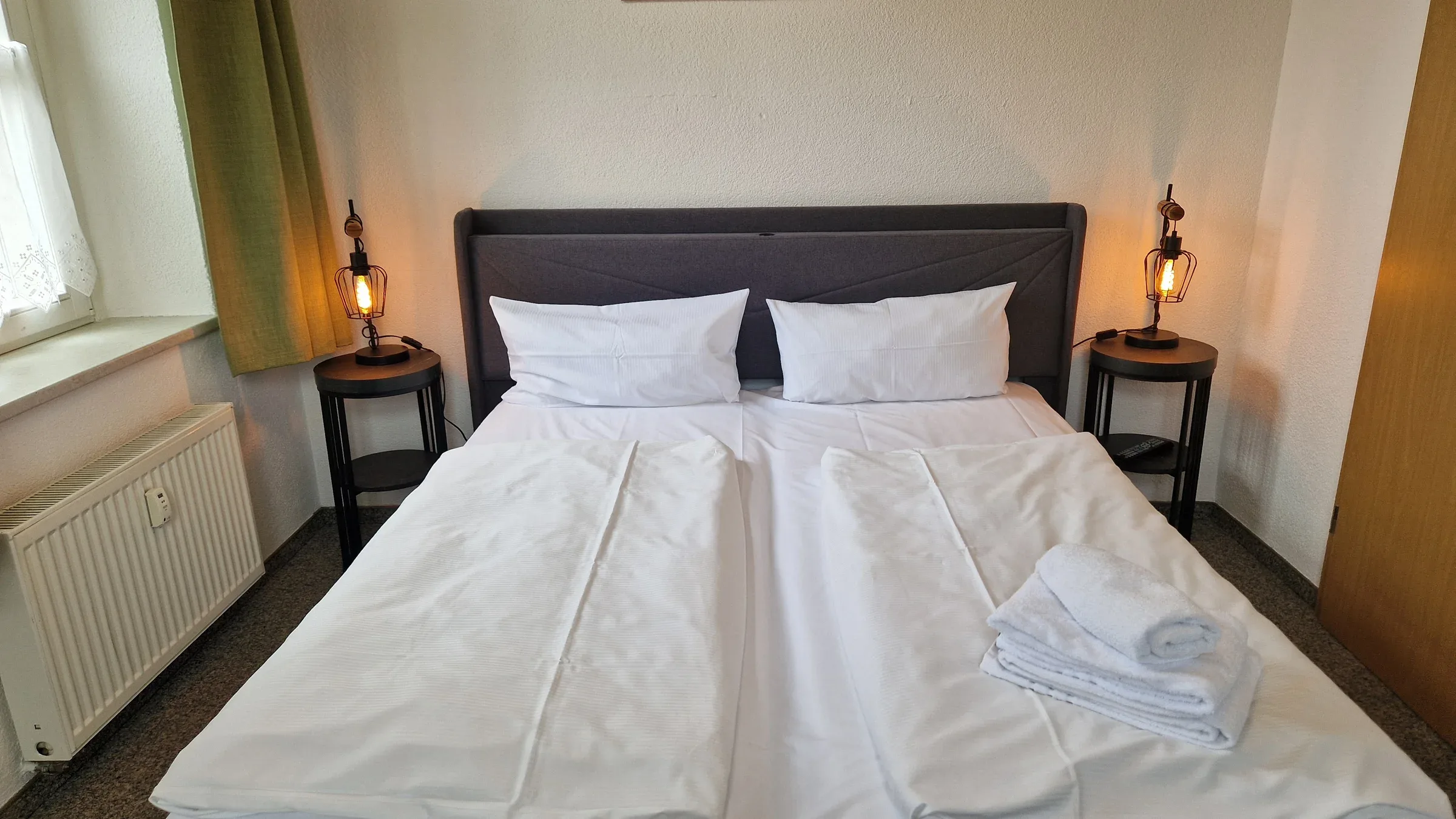 Doppelbett mit dunkelgrauem Polsterkopfteil, weißer Bettwäsche und zwei runden Nachttischen mit Industrie-Lampen