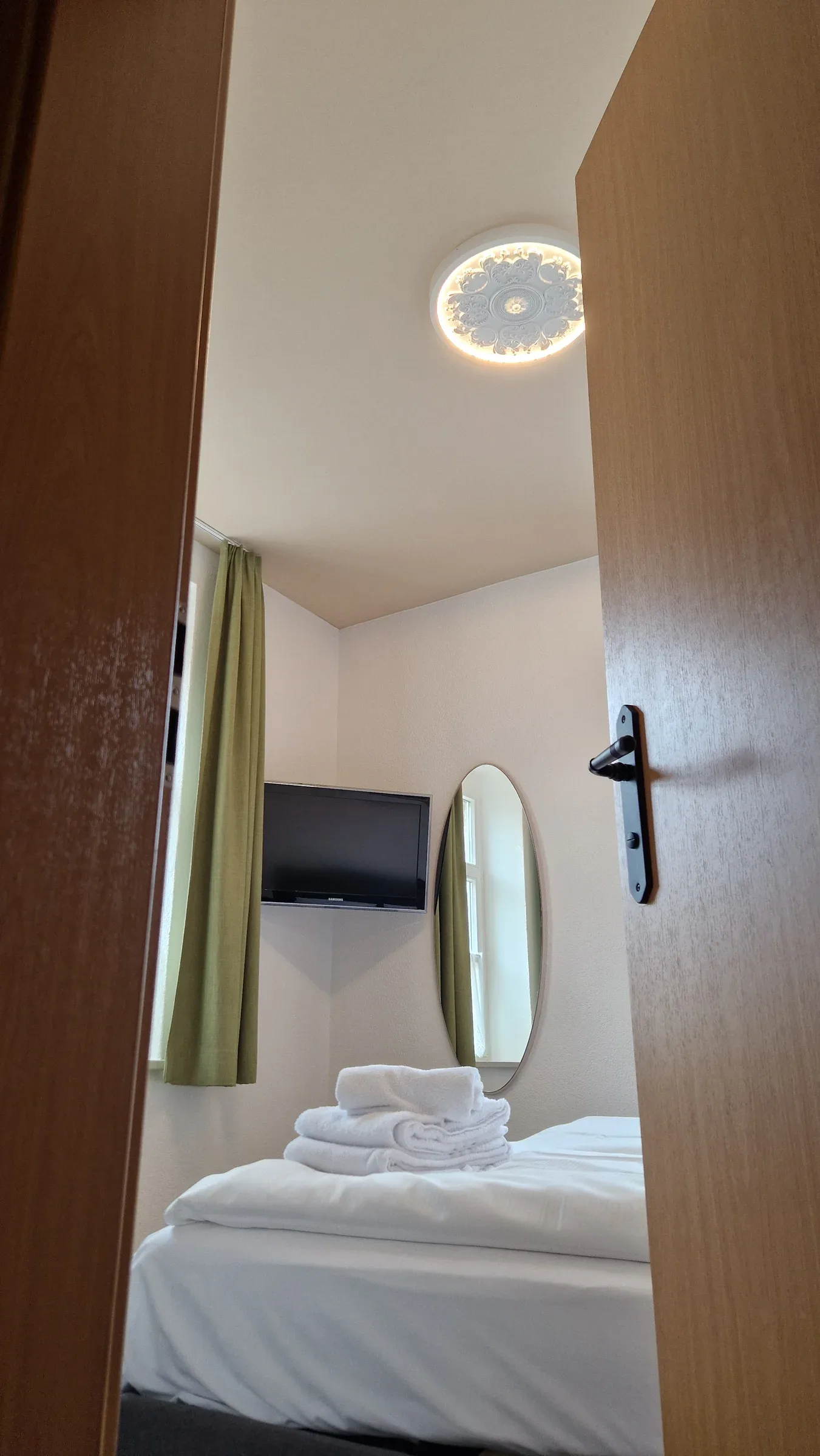 Blick durch die offene Tür ins Schlafzimmer mit Fernseher, ovalem Wandspiegel und grünen Vorhängen