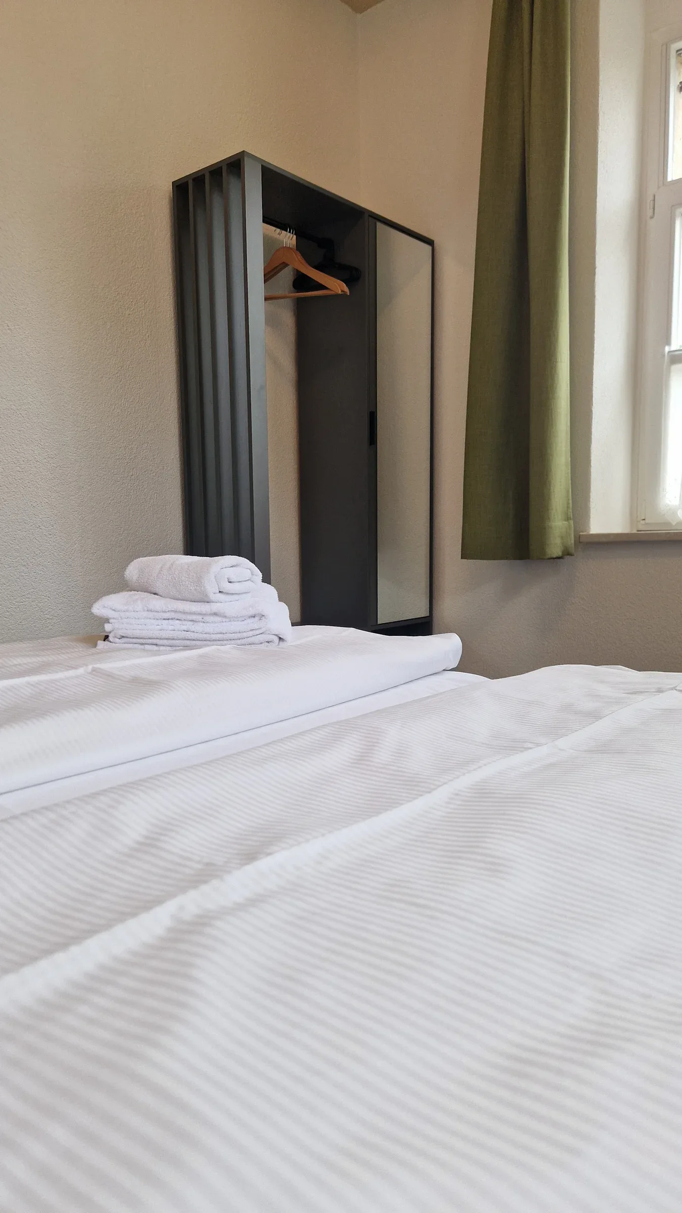 Offener Kleiderschrank mit Spiegeltür und Kleiderstange neben grünem Vorhang, gefaltete Handtücher auf dem Bett