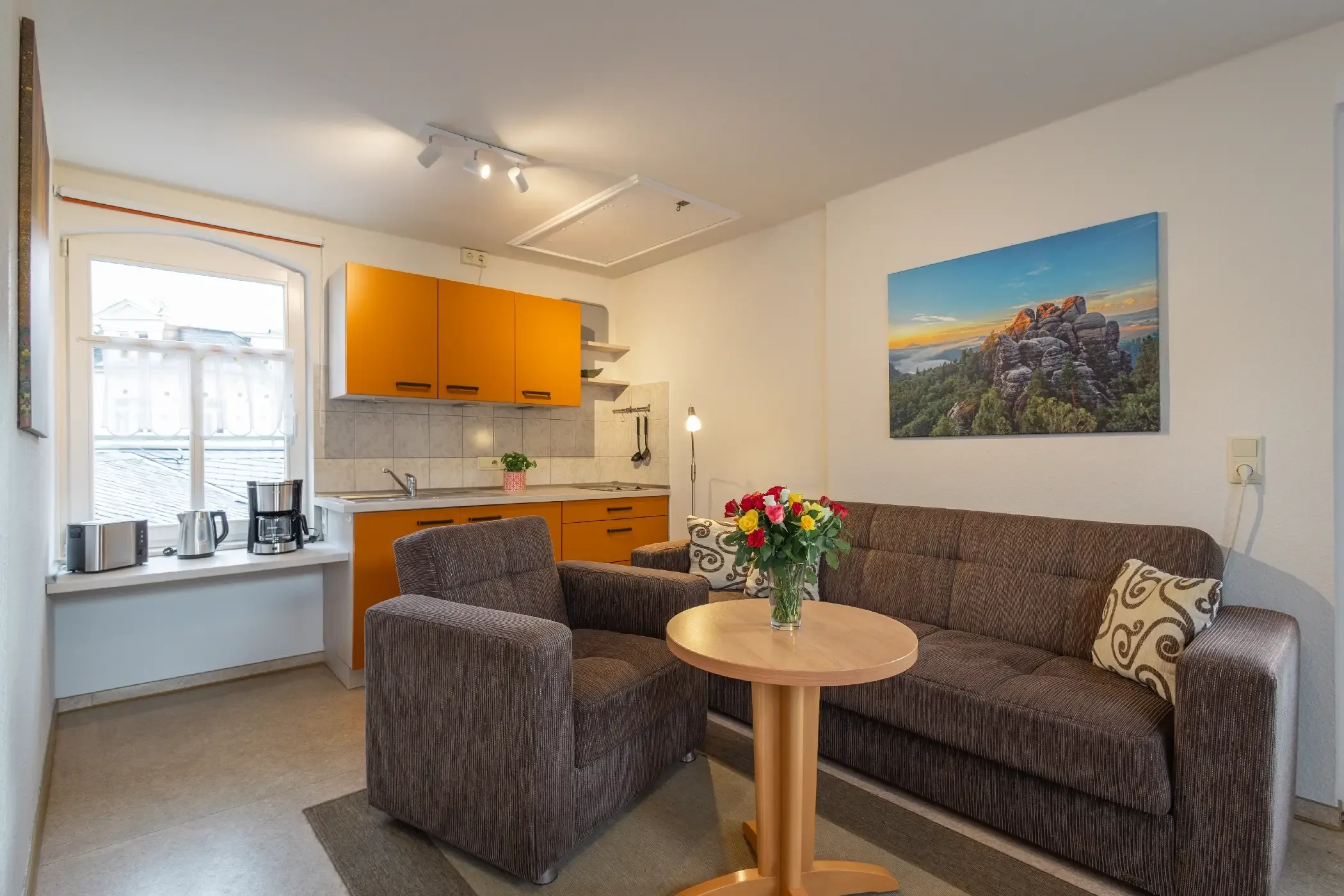 Kleine Küche mit orangen Hängeschränken neben einem Fenster und Sitzbereich mit braunem Sofa, Sessel und rundem Holztisch mit Blumenstrauß