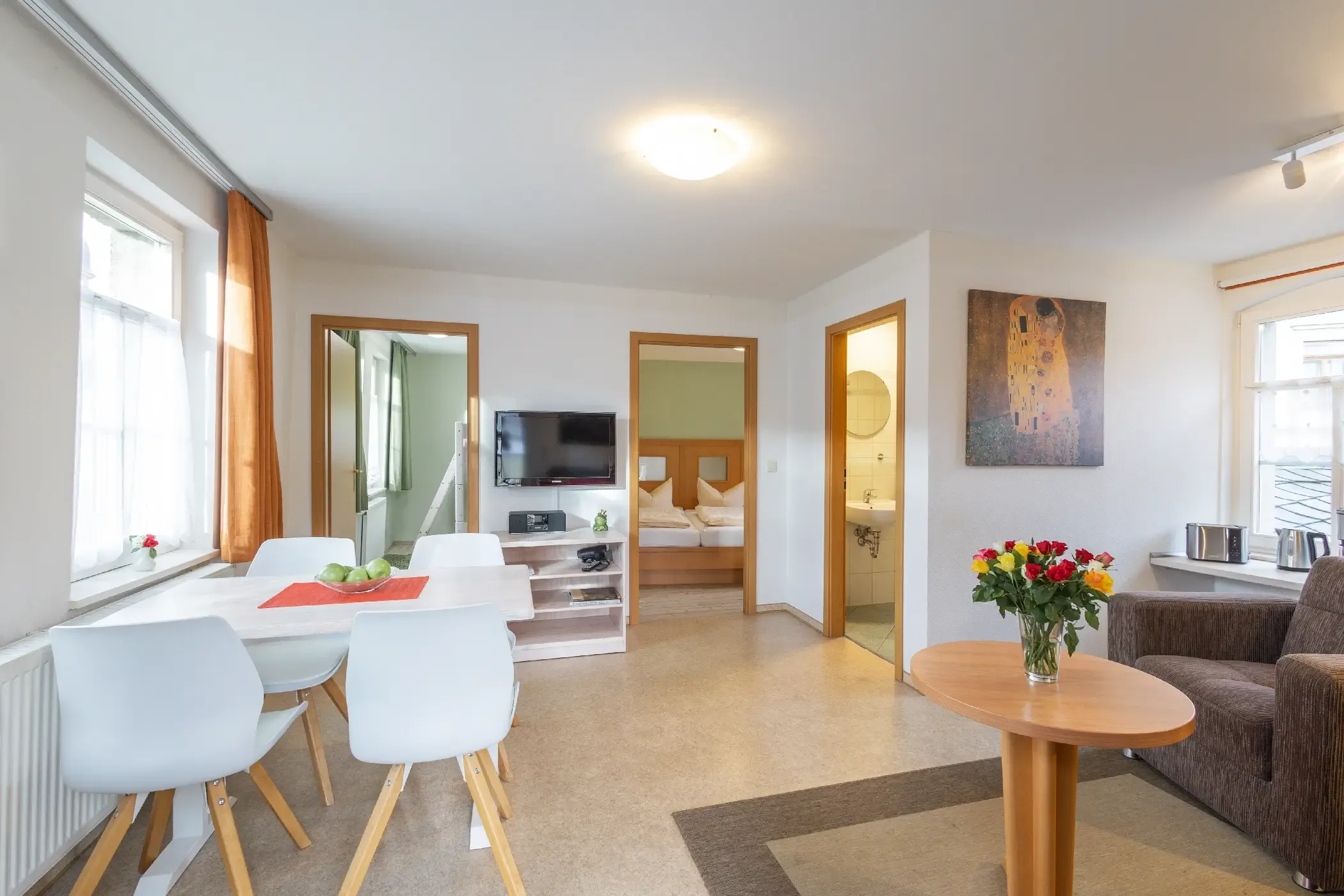 Wohnzimmer mit Esstisch, vier weißen Stühlen, Sofa, runden Holztisch mit Blumenstrauß, Fernseher an der Wand und Blick in Schlafzimmer und Bad
