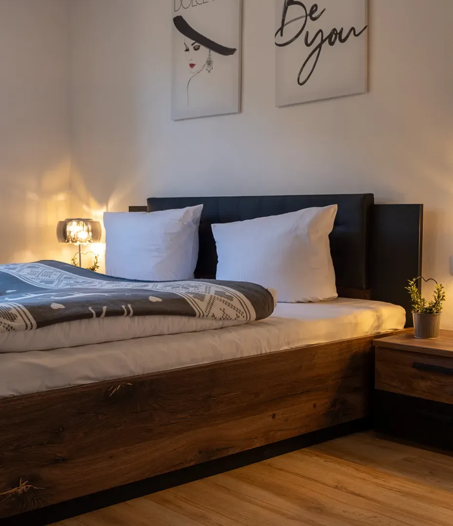 Doppelbett mit Holzbettgestell, zwei weißen Kissen, Nachttisch mit Lampe und Pflanze, zwei Wandbilder über dem Bett