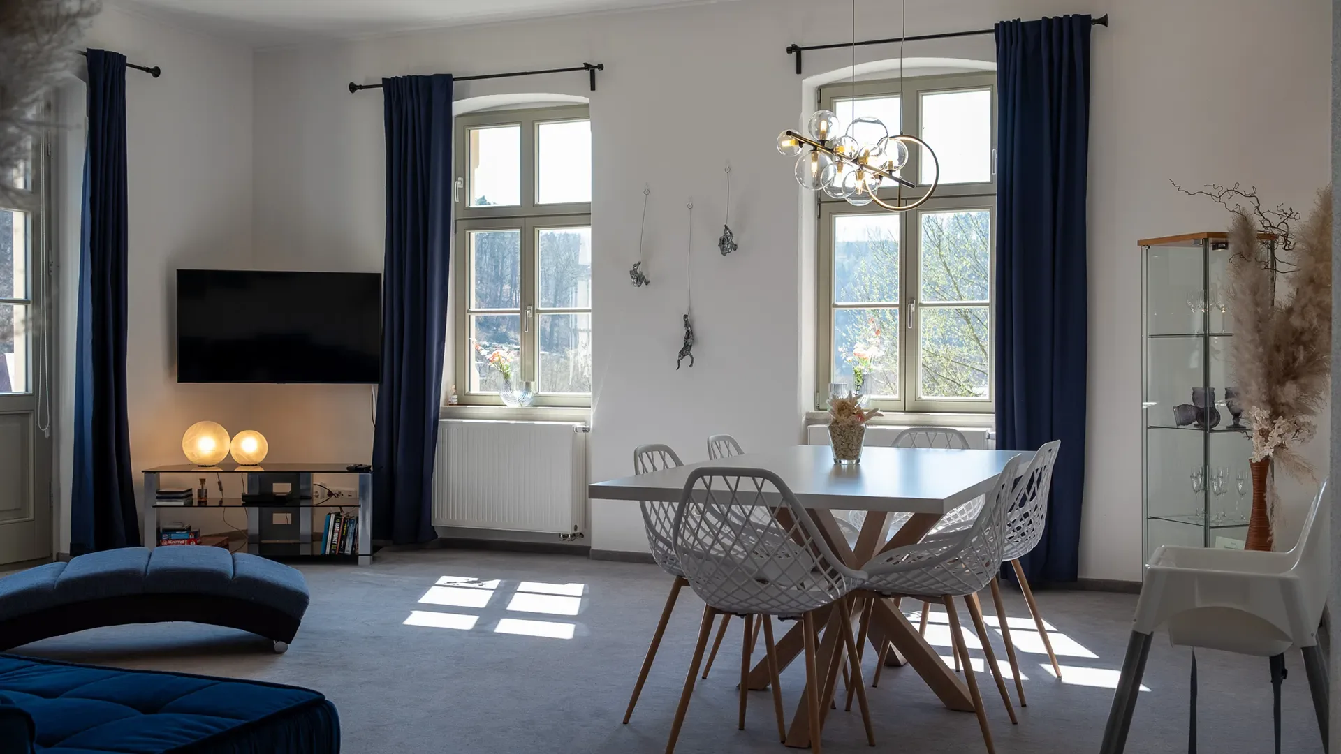 Wohnzimmer mit zwei hohen Fenstern, dunkelblauen Vorhängen, einem runden Esstisch mit vier weißen Stühlen und einem Fernseher an der Wand