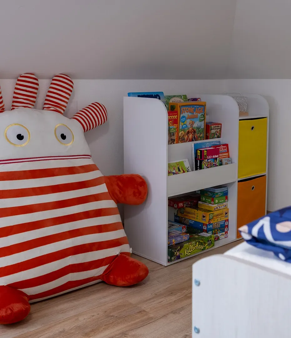 Großes Kuscheltier mit roten und weißen Streifen neben einem weißen Regal mit bunten Spielzeug- und Gesellschaftsspielen in einem Kinderzimmer