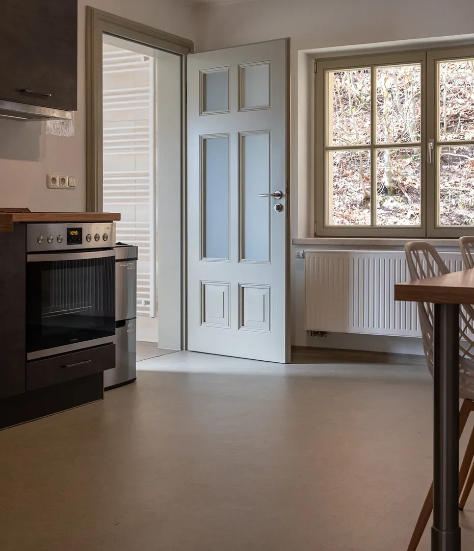 Küche mit offenem weißen Türblatt, Fenster mit Sprossen und Heizkörper darunter, links ein Backofen mit Kochfeld, rechts ein Holztisch mit Stühlen