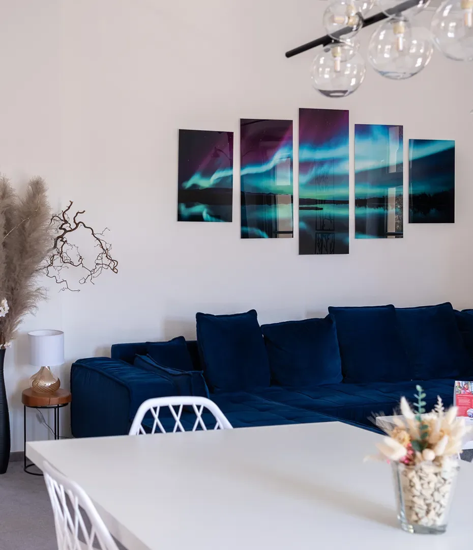 Wohnzimmer mit dunkelblauem Ecksofa, weißem Tisch mit weißen Stühlen und mehrteiliger Wandkunst mit Nordlichtmotiv