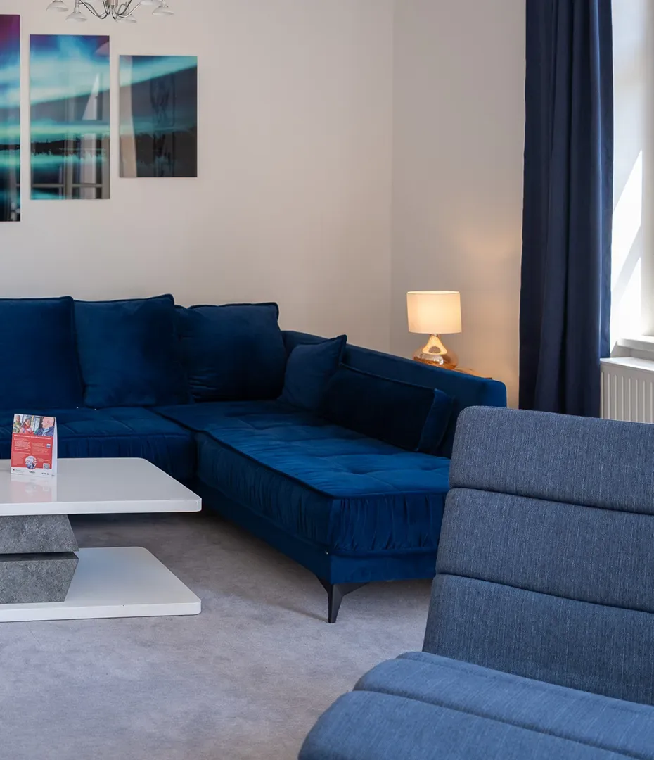 Wohnzimmer mit dunkelblauem Ecksofa, grauem Sessel, weißem Couchtisch und Tischlampe auf einem Beistelltisch vor weißer Wand