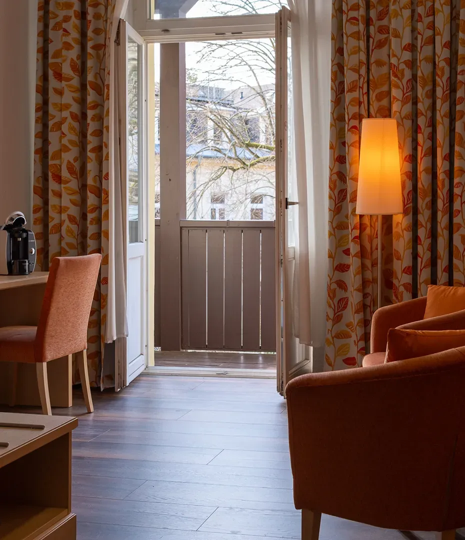 Hotelzimmer mit offenem französischem Balkon, orangefarbenen Sesseln, gemusterten Vorhängen und Stehlampe
