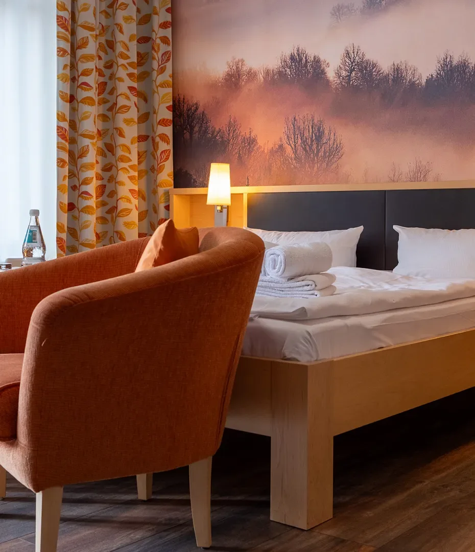 Hotelzimmer mit Einzelbett, braunem Sessel, Nachttischlampe und Wandbild mit Waldmotiv