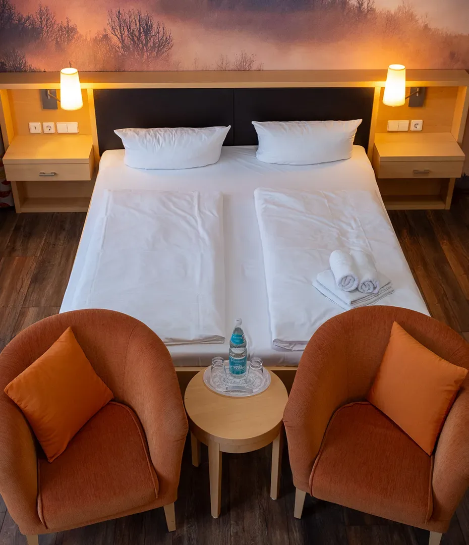Doppelbett mit weißen Kissen und Bettwäsche in einem Hotelzimmer mit zwei orangen Sesseln und kleinem runden Tisch mit Wasserflasche