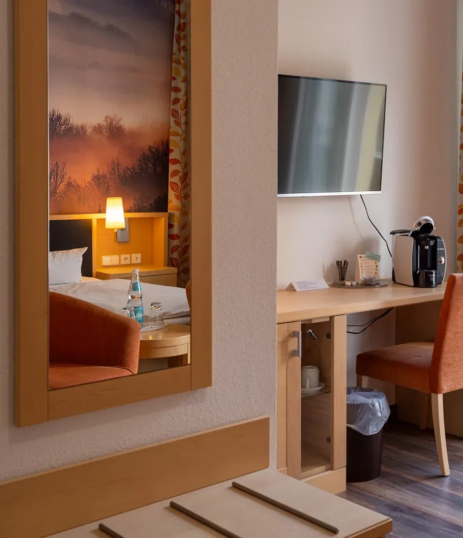Blick in ein Hotelzimmer mit Spiegel, Schreibtisch, Fernseher, Kaffeemaschine und Stuhl, im Spiegel ein Bett mit Nachttischlampe und Wandbild