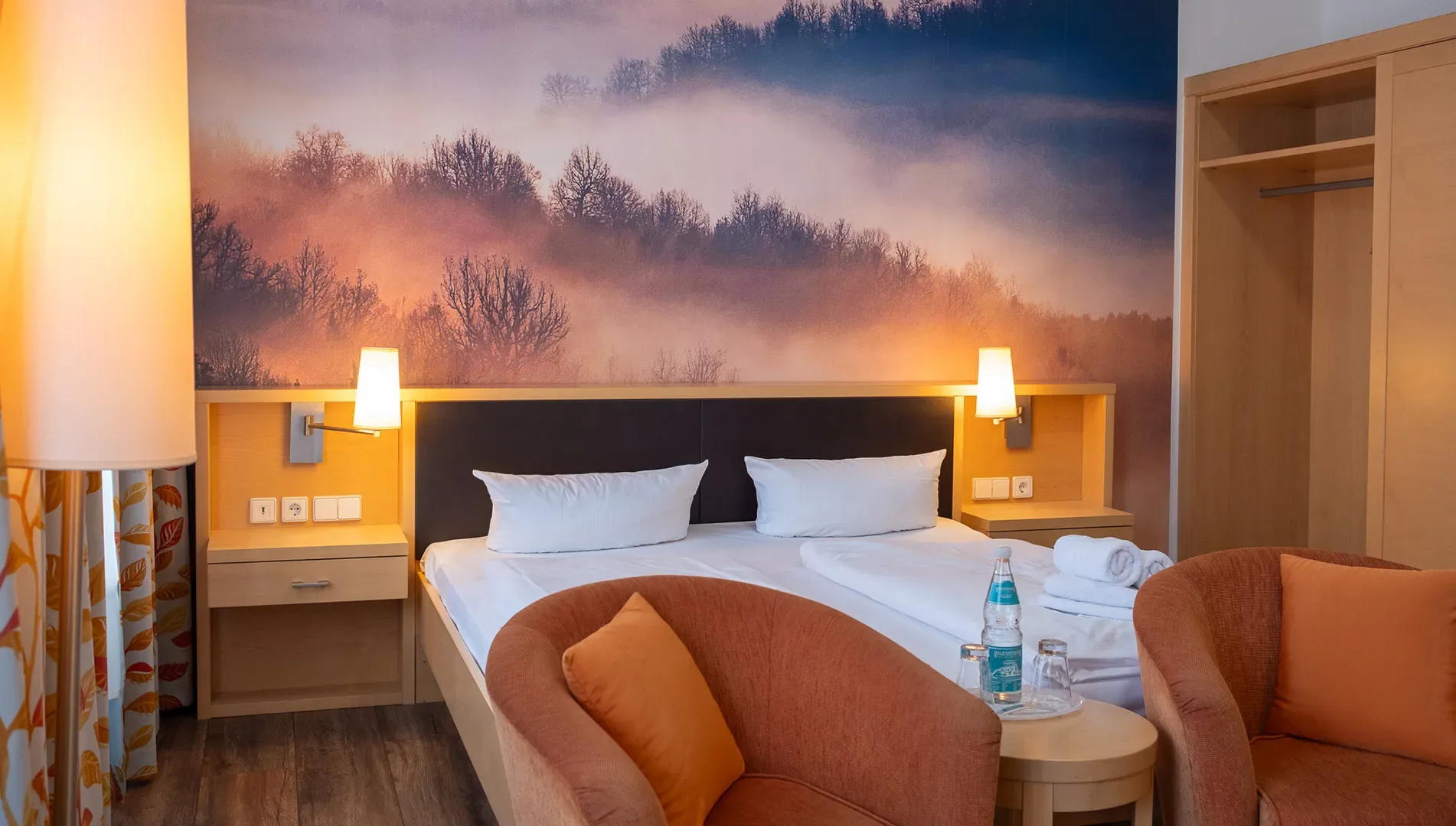 Hotelzimmer mit Doppelbett, zwei Nachttischen mit Wandlampen, zwei Sesseln mit Kissen, kleinem Tisch mit Wasserflasche und Gläsern sowie offener Garderobe
