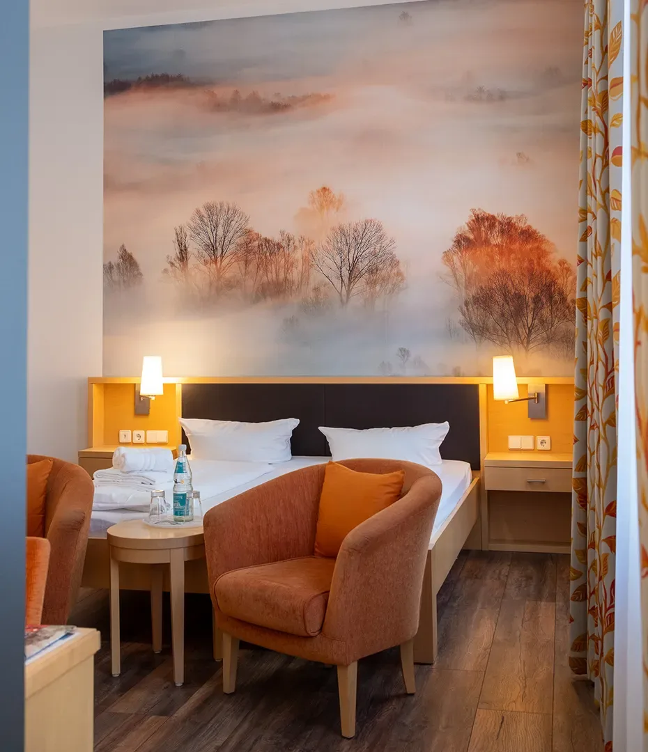 Hotelzimmer mit Doppelbett, zwei orangen Sesseln, kleinem runden Tisch und großem Wandbild mit nebliger Landschaft und Bäumen